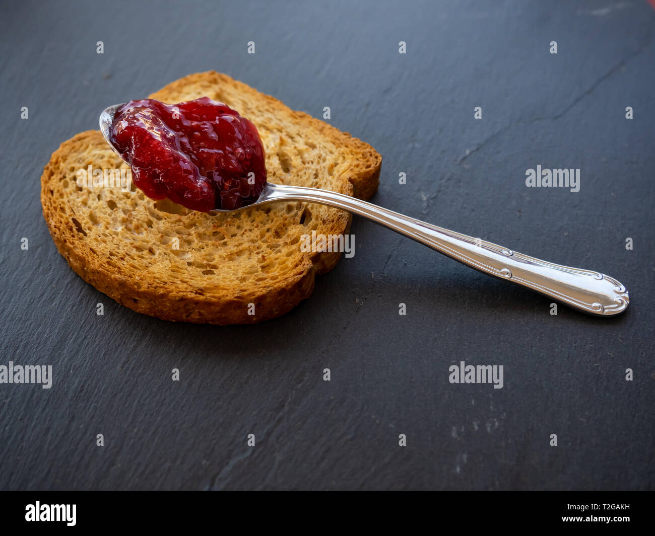 A spoon with raspberry jam on a slice of toast on a plate slate Stock Photo