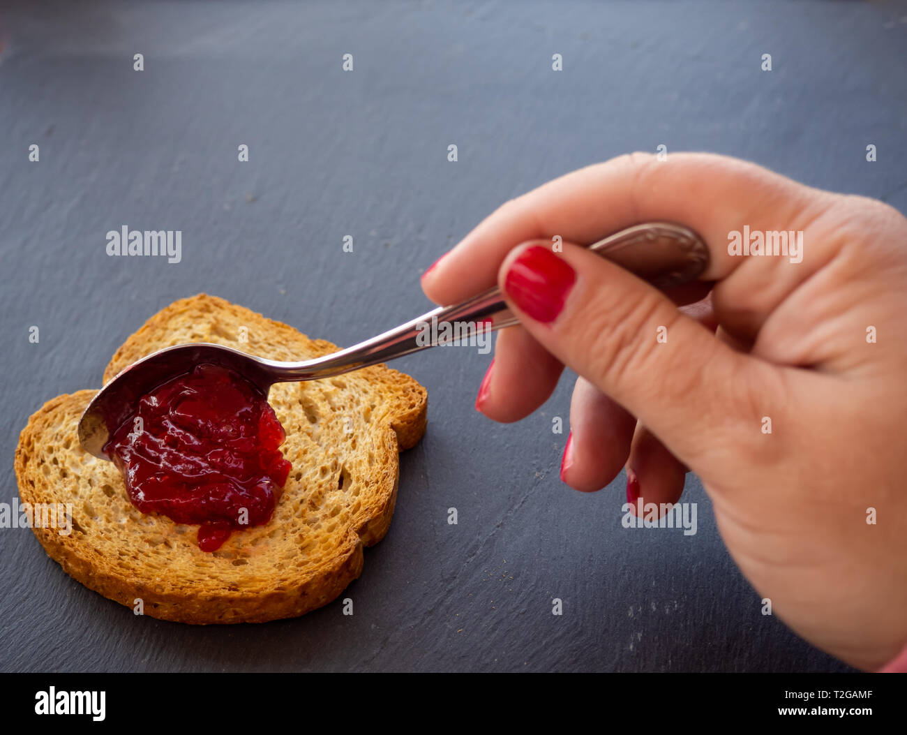 A woman with painted nails smearing red raspberry jam with a spoon on a slice of toast Stock Photo