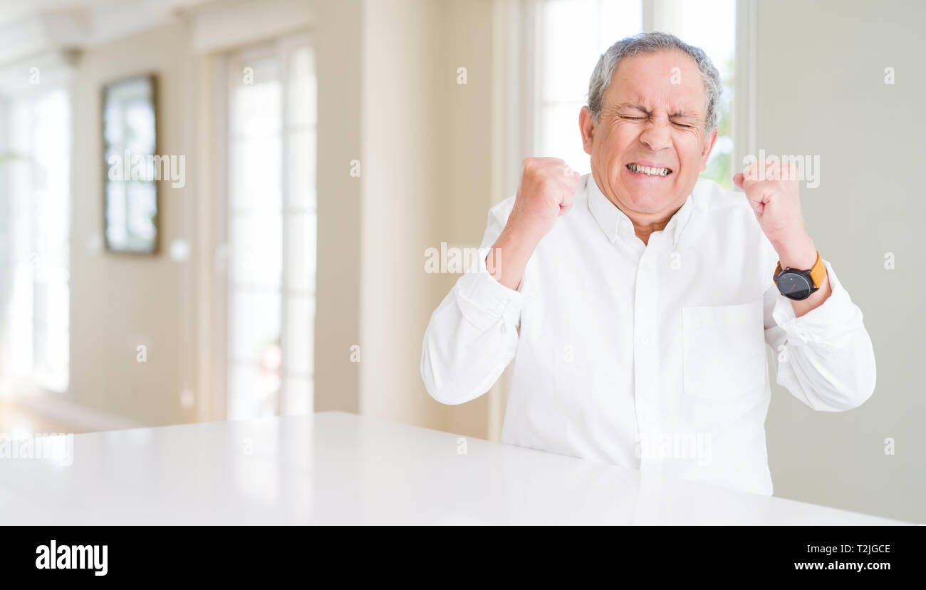 Handsome senior man at home excited for success with arms raised ...