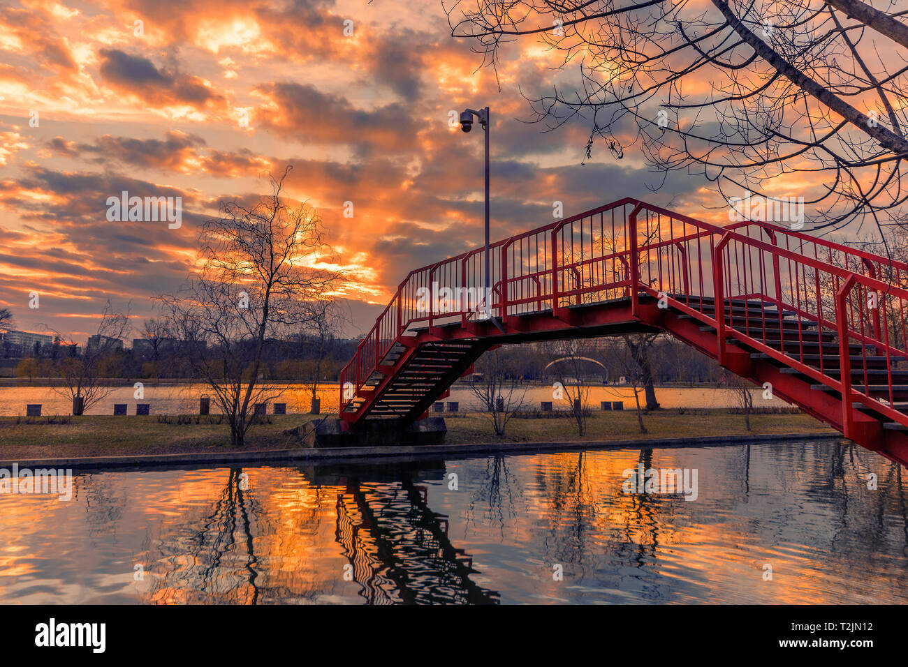 Sunset seen from a park in Bucharest with sun rays coming out from the clouds and a red metal bridge in the foreground shot at the beginning of the sp Stock Photo