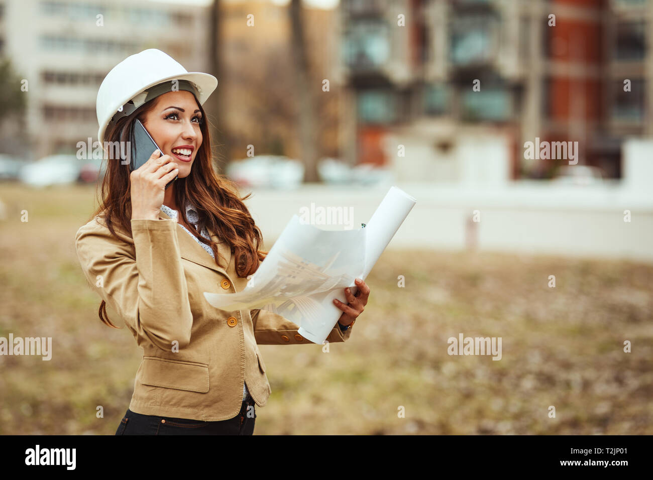 Young successful female engineer with white helmet and blueprints in her hands at construction site analyzing new project and talking on a smartphone  Stock Photo