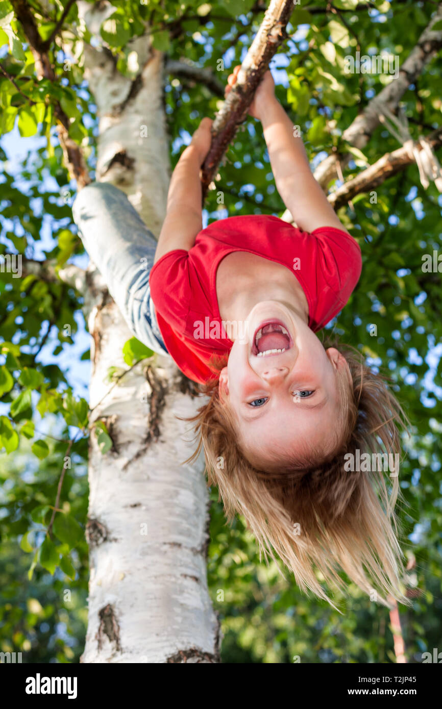 Portrait of happy elementary age girl wearing red tee shirt hanging upside down from a tree branch - summer fun concept Stock Photo