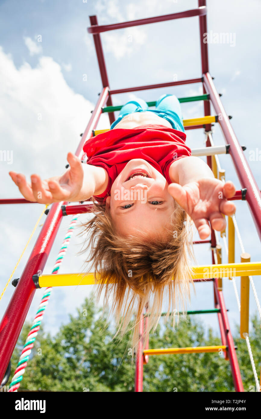 Elementary age girl hanging upside down from a jungle gym (monkey bars or climbing frame) in a playground enjoying summertime Stock Photo