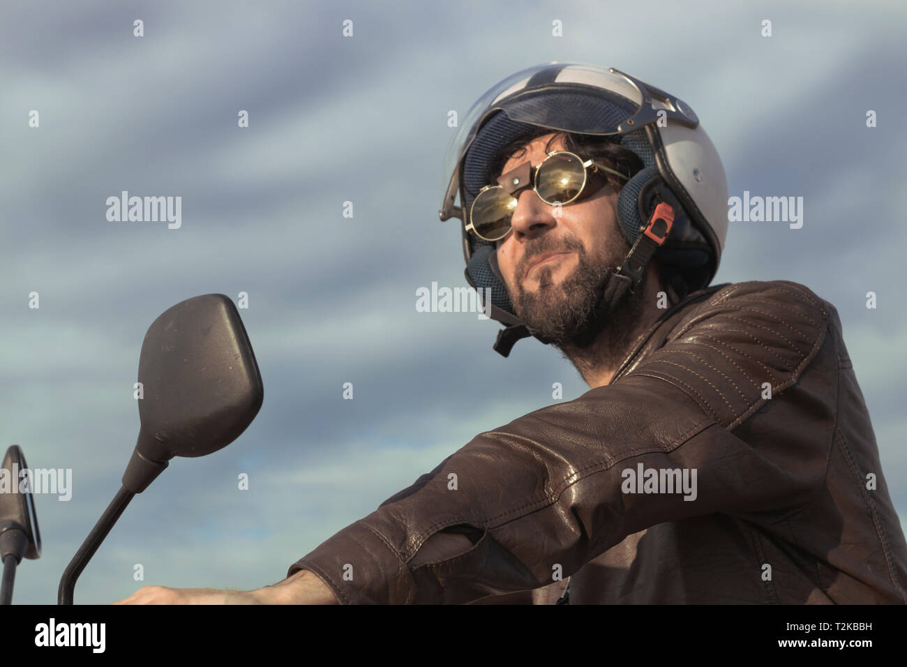 Brunette man with brown leather jacket, helmet and glasses on a retro style motorbike Stock Photo