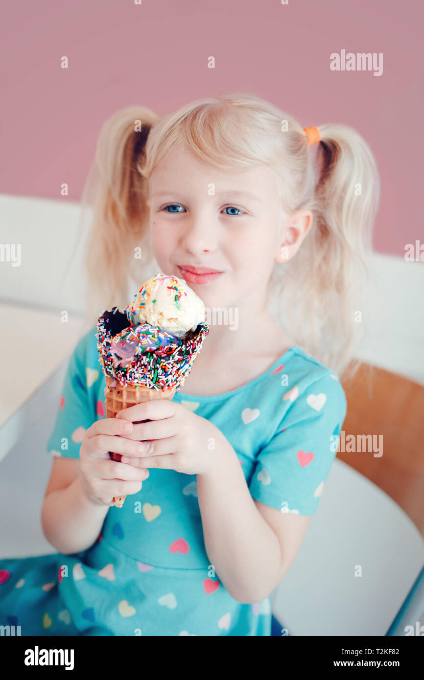 Cute adorable funny Caucasian blonde preschool girl child with blue eyes and pigtails eating licking ice cream in large waffle cone with sprinkles. Ha Stock Photo
