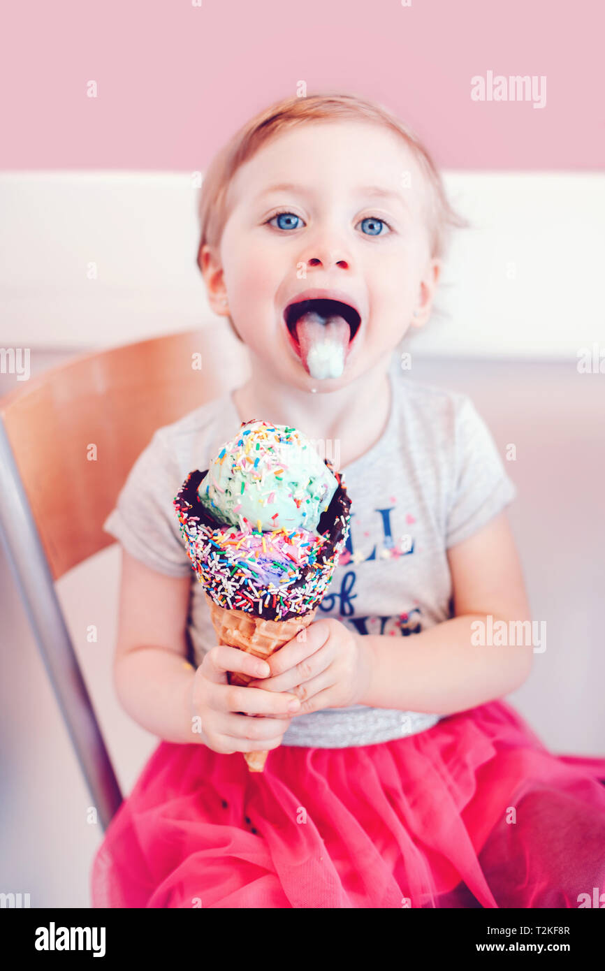 Cute funny Caucasian blonde babyl girl child with blue eyes eating licking ice cream in large waffle cone with sprinkles and showing tongue. Happy chi Stock Photo