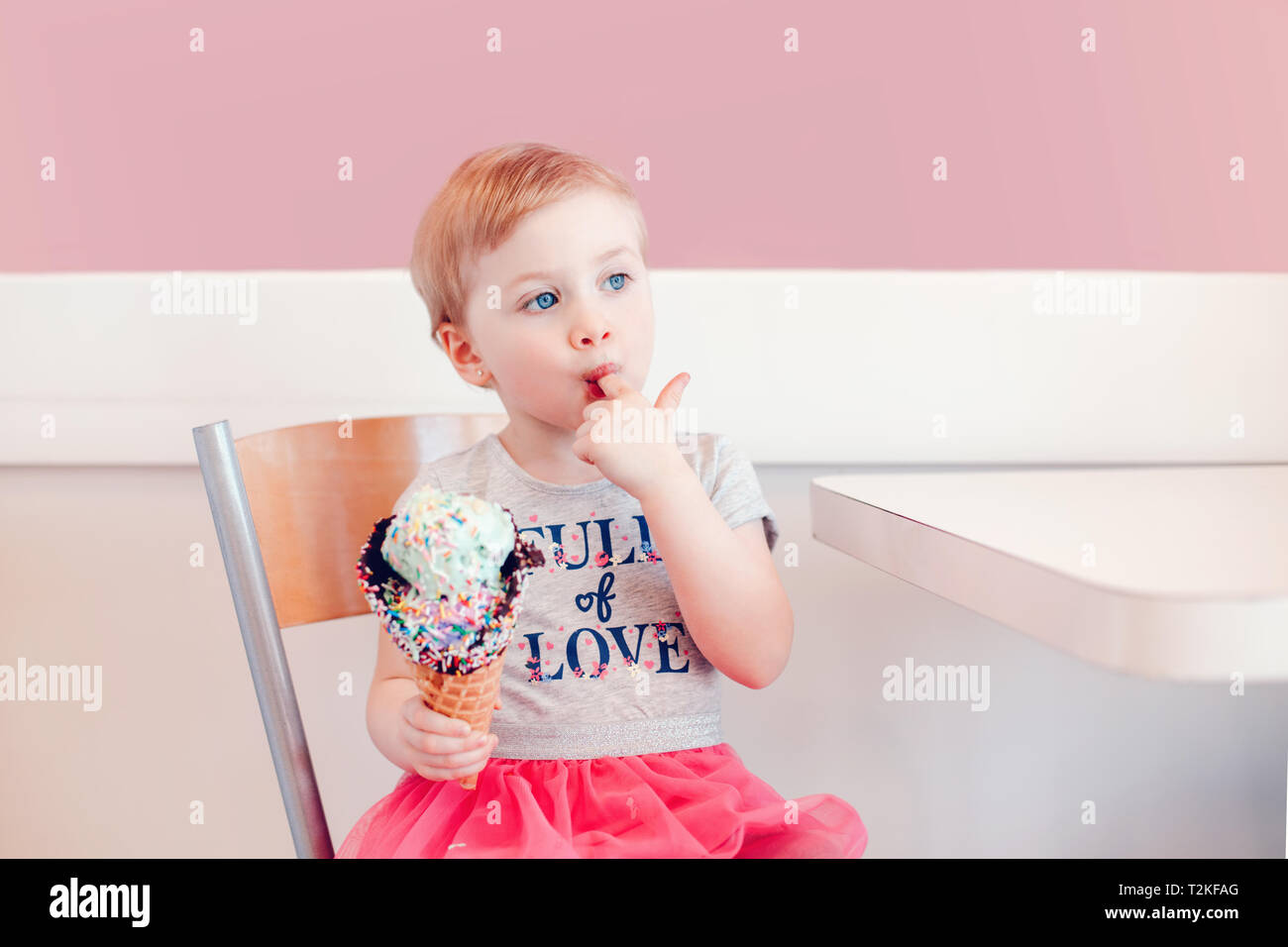 Cute adorable funny Caucasian blonde babyl girl child with blue eyes eating ice cream in large waffle cone with colorful sprinkles. Kid licking finger Stock Photo