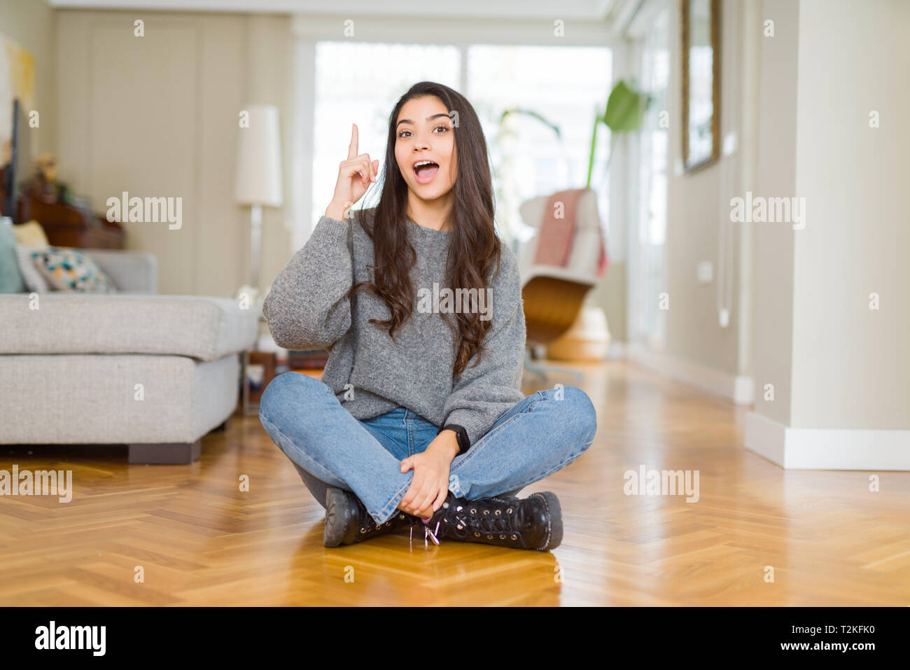 Young beautiful woman sitting on the floor at home pointing finger up ...