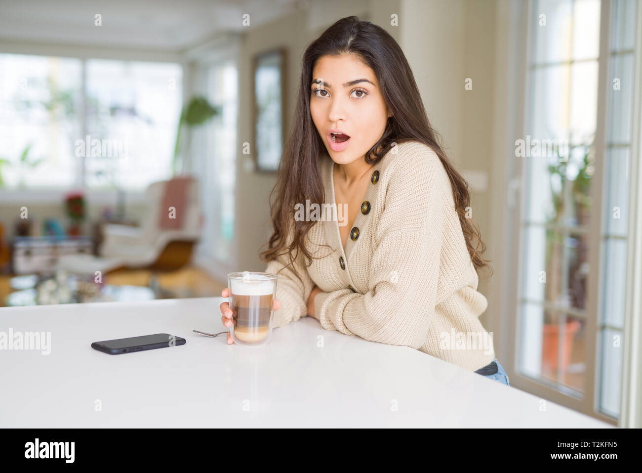 Young woman drinking a cup of coffee at home scared in shock with a