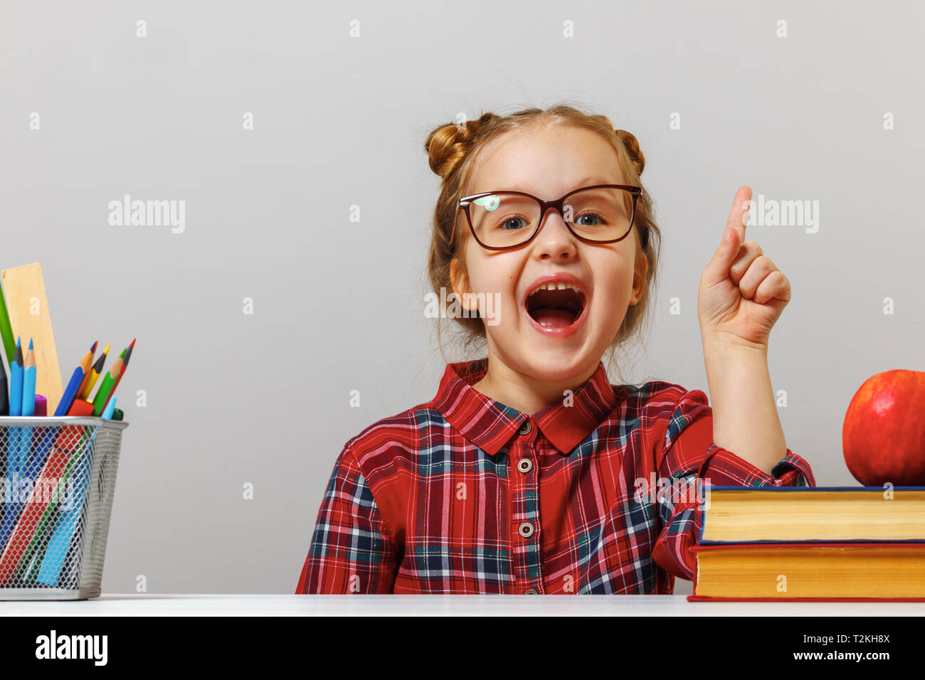 Funny little preschool girl in glasses sits at the table with books and points finger up. The concept of education. Gray background. Stock Photo