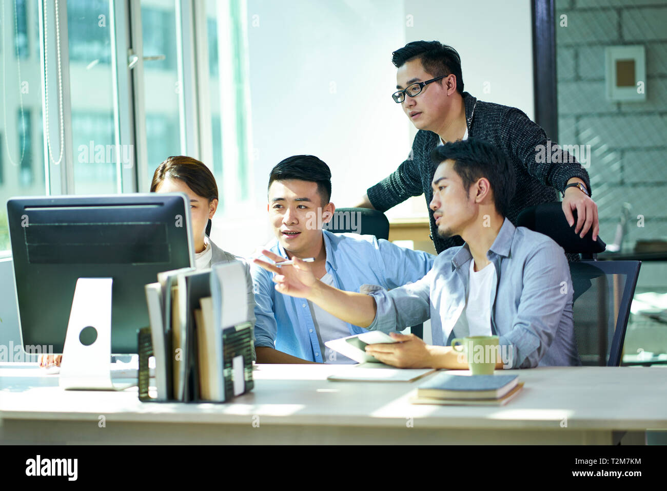 four young asian entrepreneurs working together discussing business plan using desktop computer in office. Stock Photo