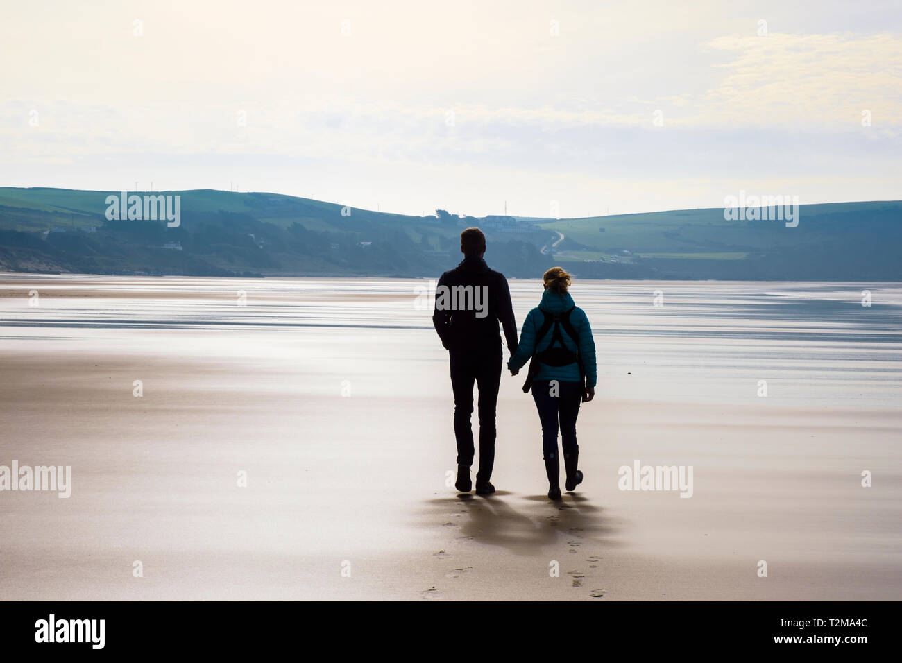 A couple holding hands walking together on a quiet sandy beach at low tide. Woolacombe, North Devon, England, UK, Britain Stock Photo