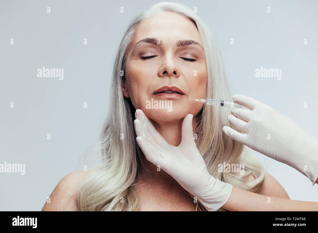 Senior woman having skin injections against grey background. Female getting anti aging shots on her face by beautician. Stock Photo