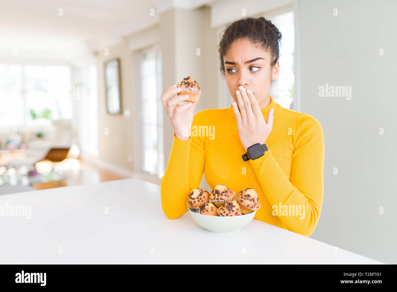 Young african american woman eating chocolate chips muffins cover mouth with hand shocked with shame for mistake, expression of fear, scared in silenc Stock Photo