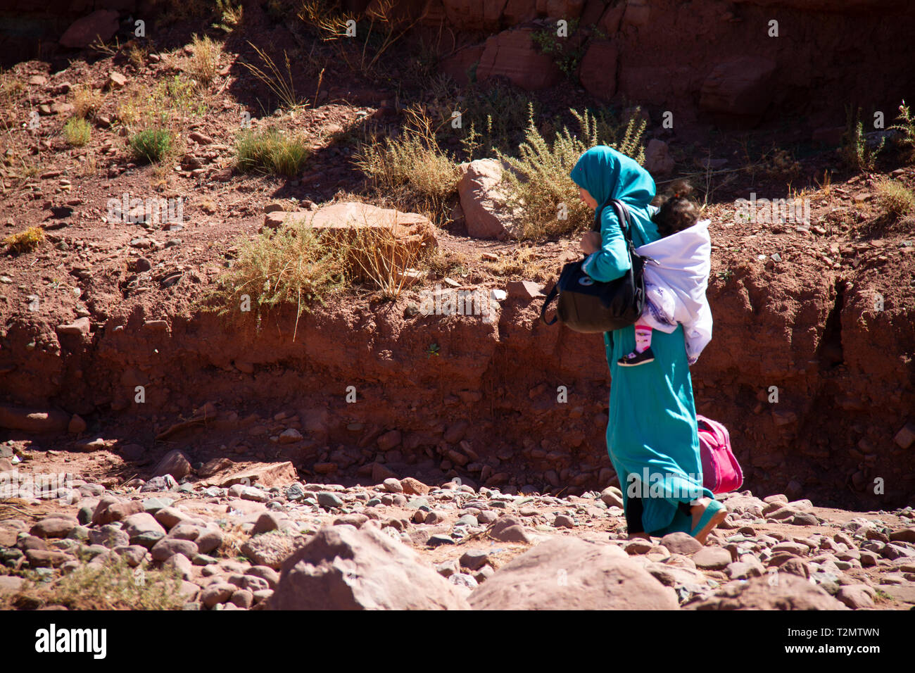 Morocco Local Bedouin Mother, in Turquoise Robe, Carrying Child on back Walking in Wadi near Telouet Kasbah Stock Photo
