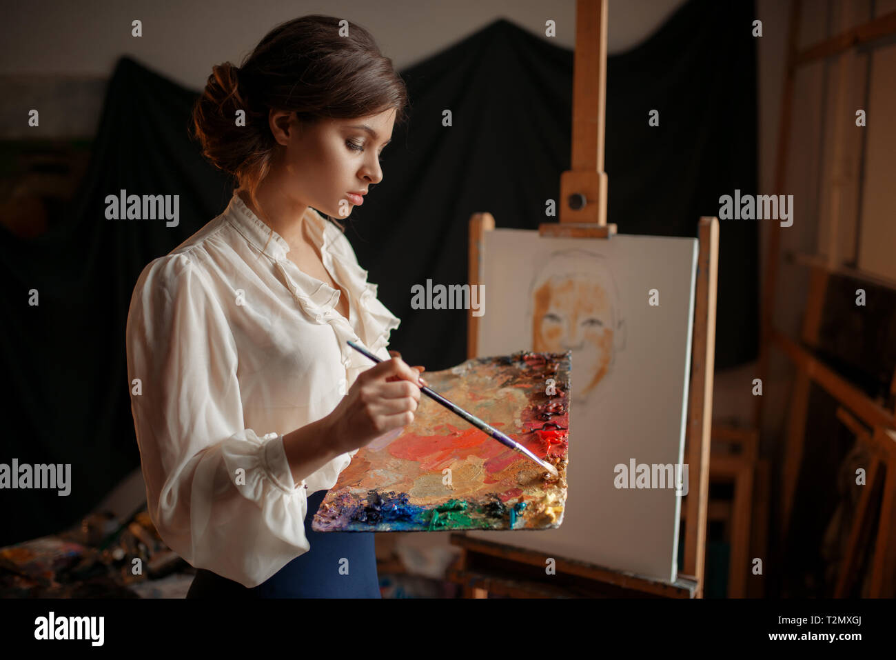 Female artist with color palette and brush standing against easel in ...