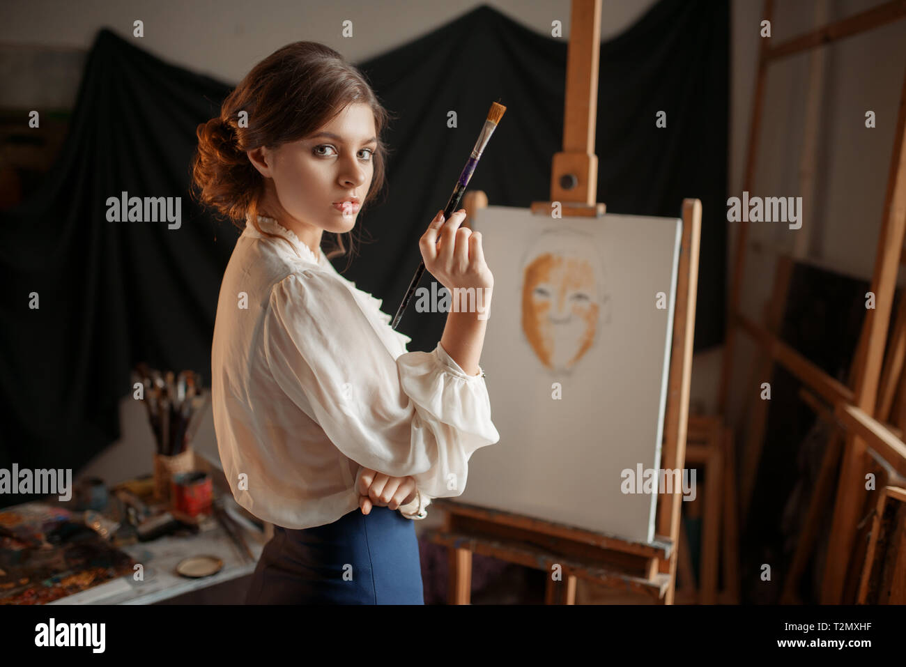 Female artist with color palette and brush standing against easel in ...