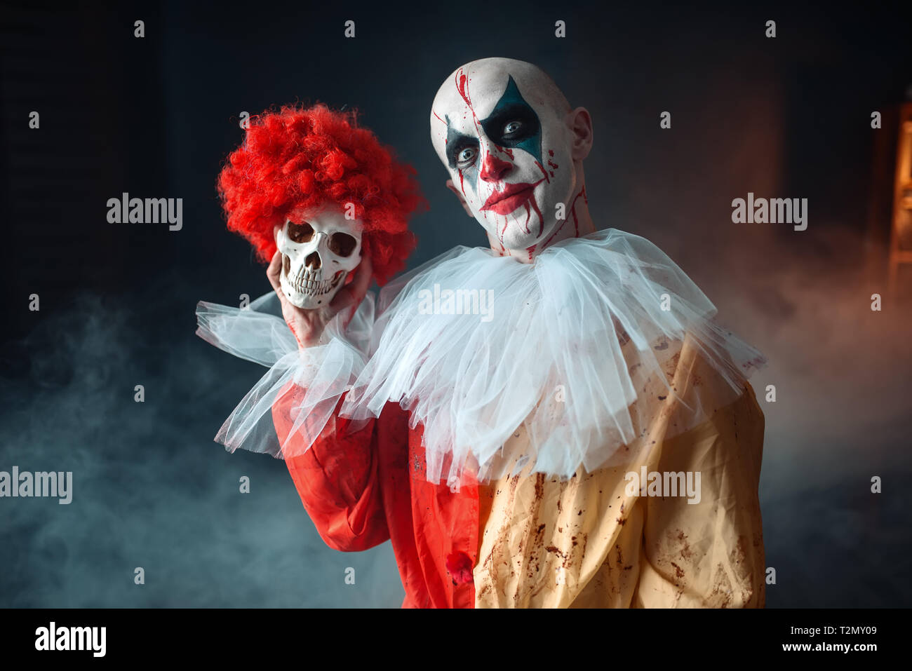 Crazy bloody clown holds human skull in red wig horror. Man with