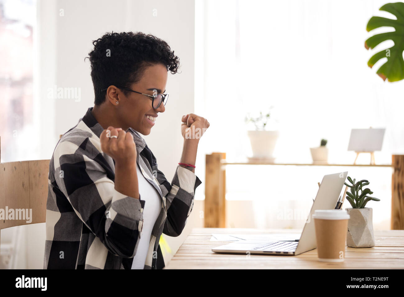 Happy black girl sitting at the desk looking at laptop. Excited woman feels happy received a scholarship, took on a good post or getting reward. Posit Stock Photo