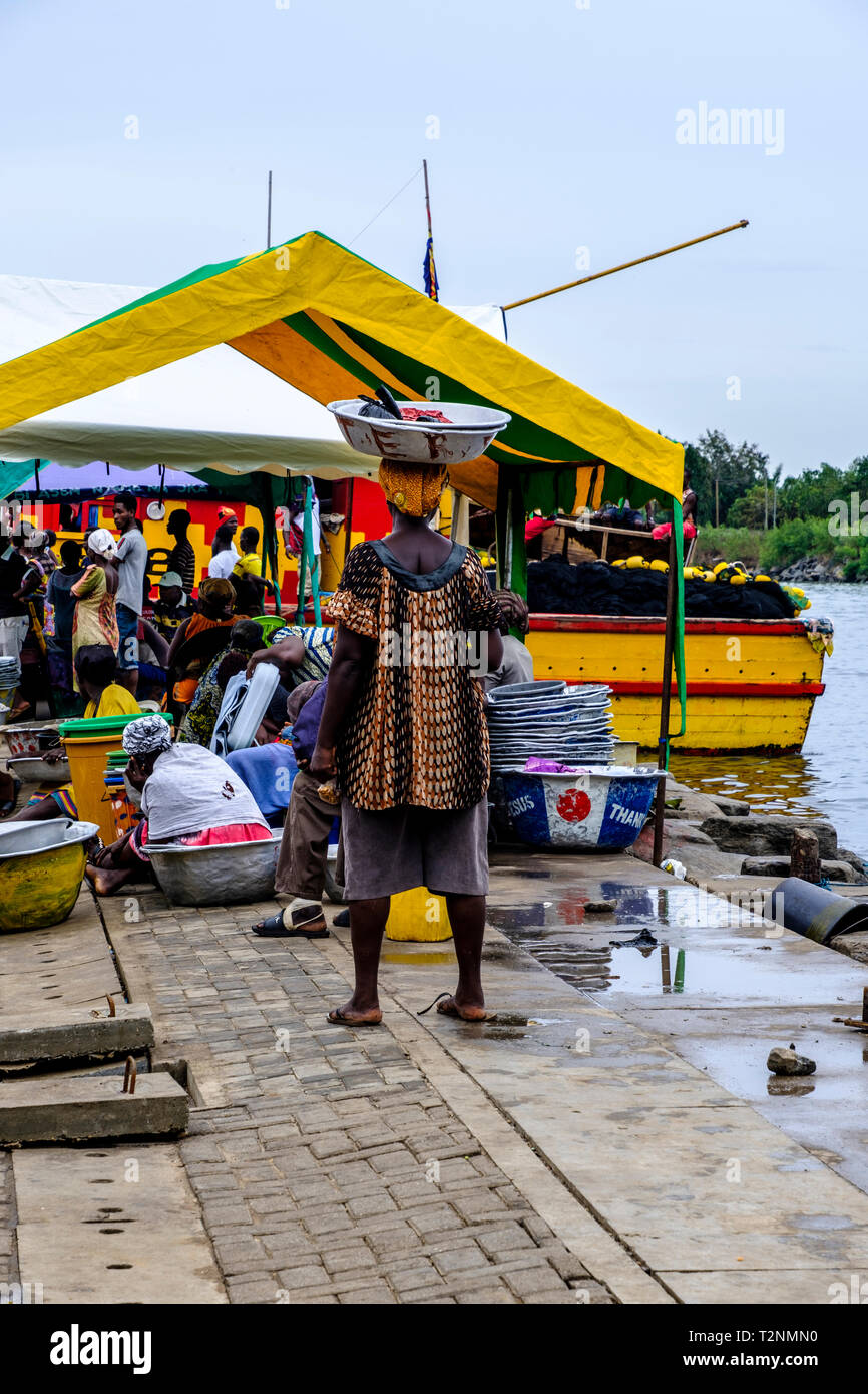 SEKONDI TAKORADI, GHANA – 10 APRIL 2018: Vibrant harbor fishing market scene as woman in colorful dress carries metal rustic bucket on her head ready  Stock Photo