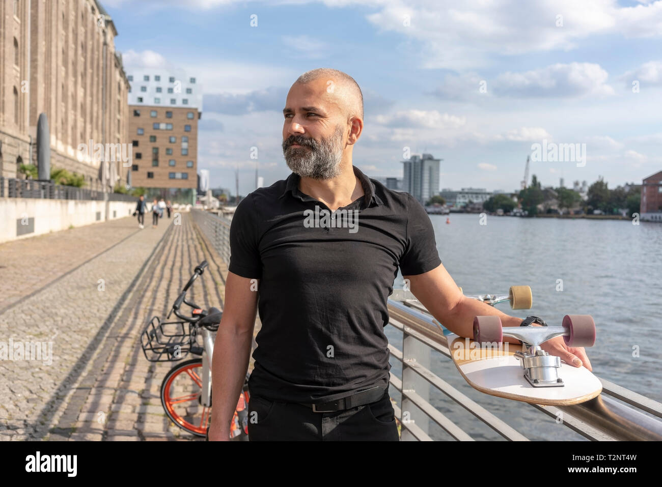 Man holding skateboard on bridge, river and buildings in background, Berlin, Germany Stock Photo
