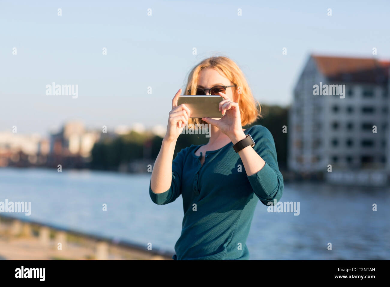 Young woman taking photograph with smartphone by river in summer, Berlin, Germany Stock Photo