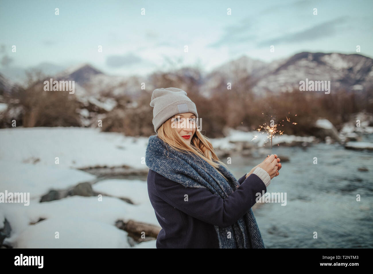 Woman in knitted hat holding sparkler on snow covered riverbank, portrait,  Orta, Piemonte, Italy Stock Photo