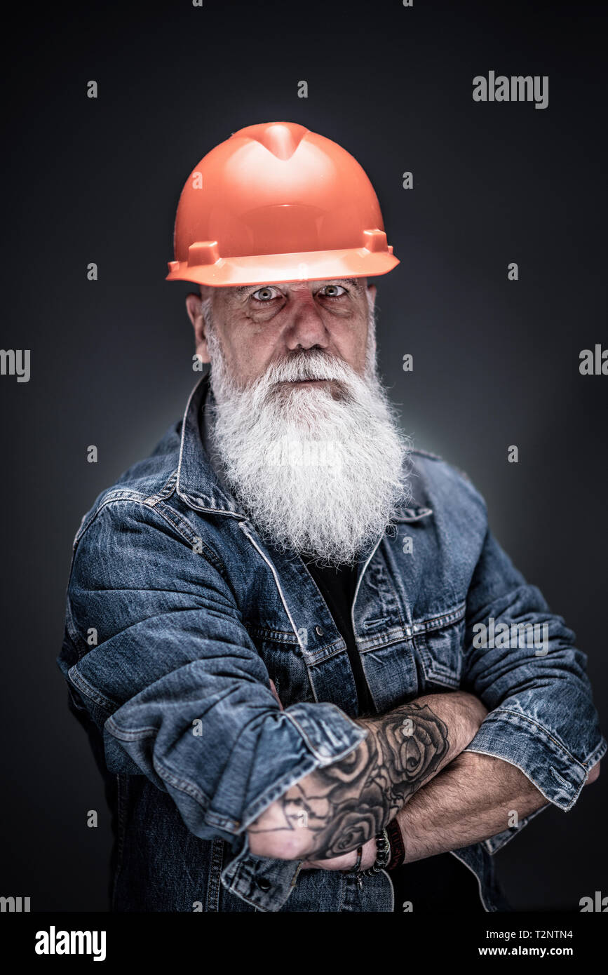 studio portrait of a senior hipster with tattooed arms, wearing a construction helmet and a long white beard Stock Photo
