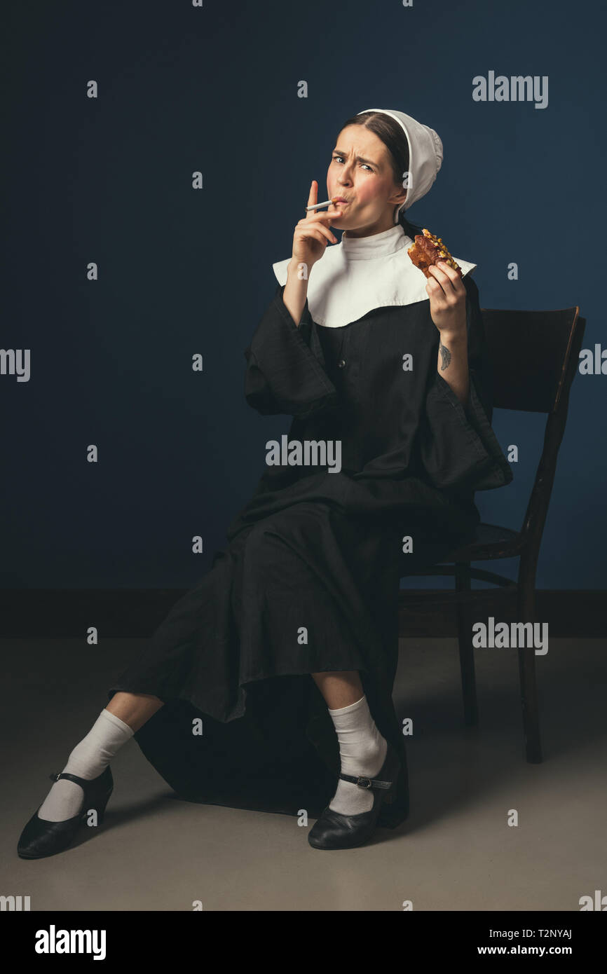 Breaking bad. Medieval young woman as a nun in vintage clothing sitting on  the chair on dark blue background. Smoking cigarette and eating hot dog in  Stock Photo