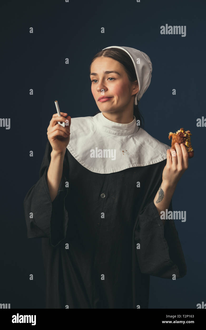 cigarette hot dog No more problems. Medieval young woman as a nun in vintage clothing sitting  on the chair on dark blue background. Smoking cigarette and eating hot dog  Stock Photo