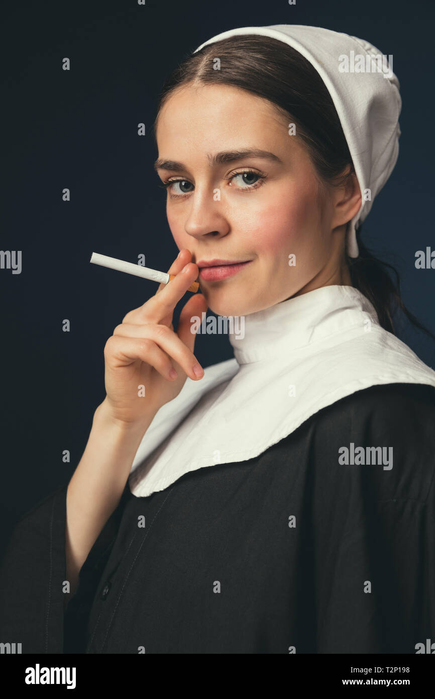 Rebellious youth. Medieval young woman as a nun in vintage clothing and white mutch with long hair standing on dark blue background. Smoking cigarette Stock Photo