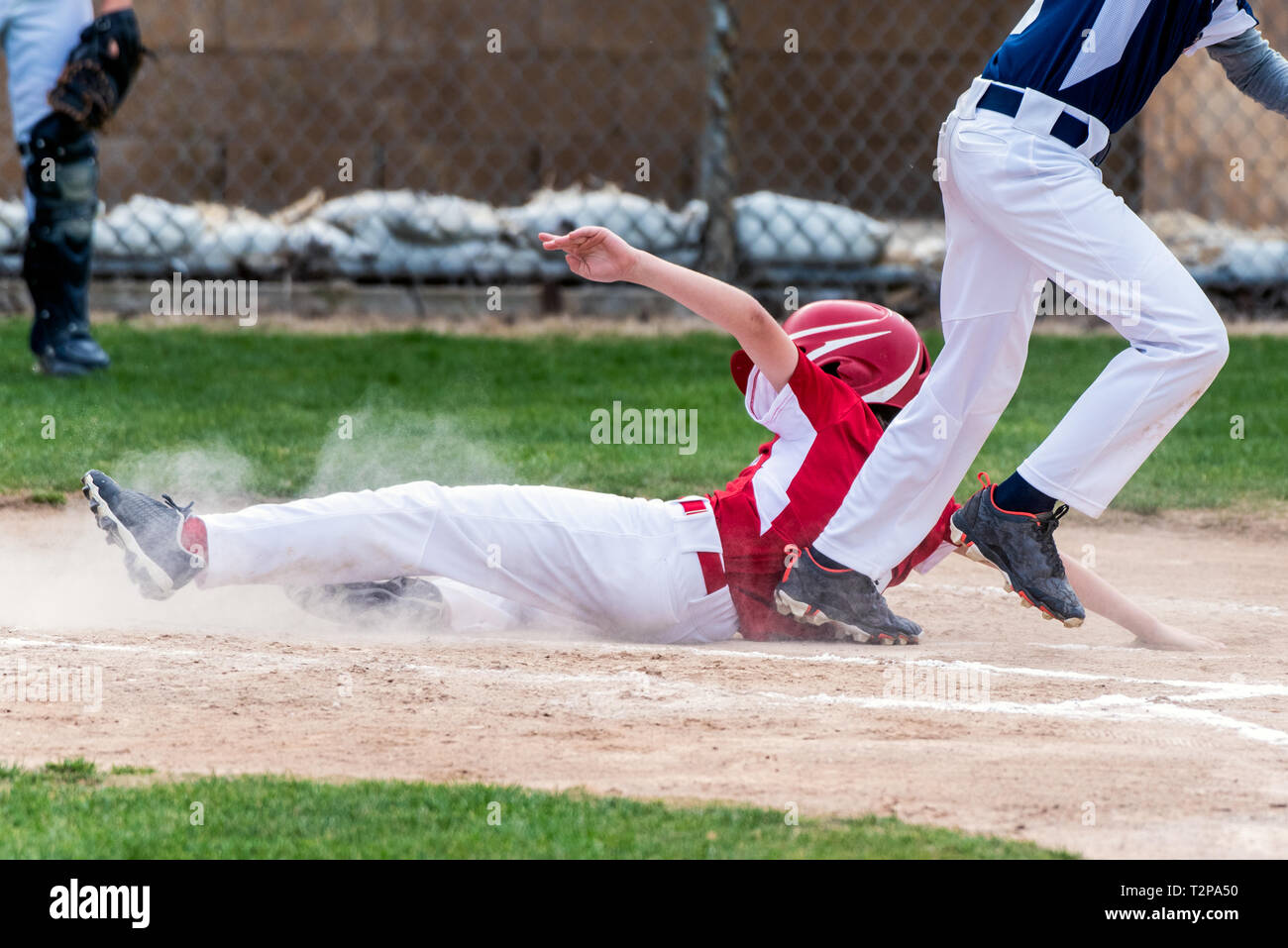 Youth baseball player in red uniform sliding safely into home plate in a cloud of dust during a game. Stock Photo