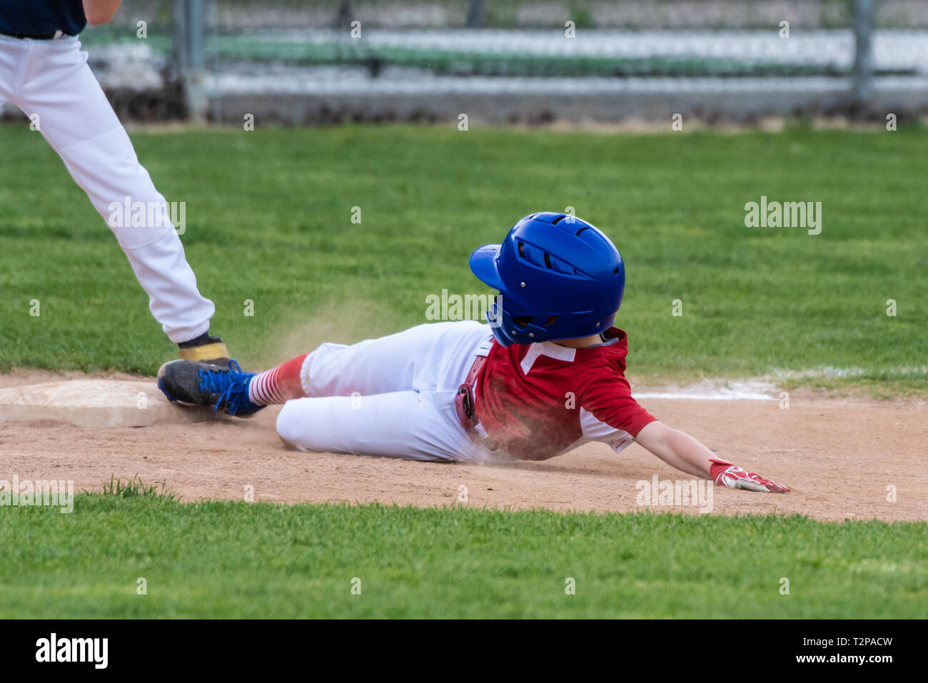 Youth baseball player in red uniform sliding safely into third base in a cloud of dust during a game. Stock Photo
