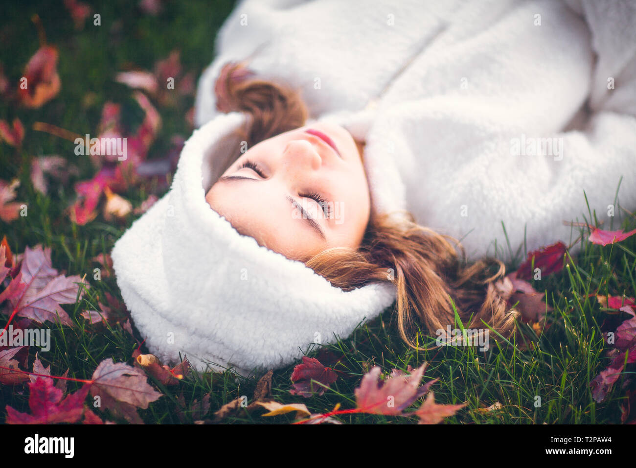 Girl in white hooded top with eyes closed, lying on grass amongst autumn leaves, portrait Stock Photo