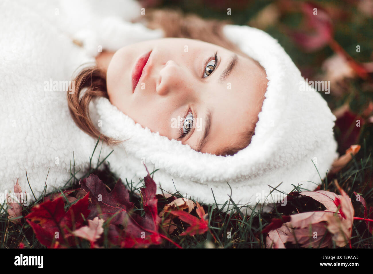 Girl in white hooded top lying on grass amongst autumn leaves, portrait Stock Photo