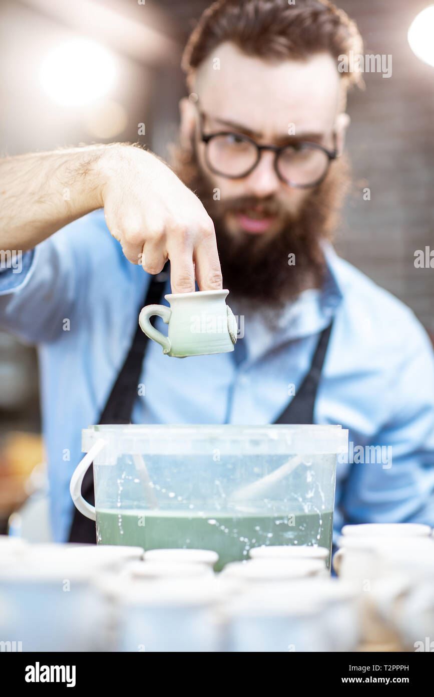 Handsome creative bearded worker painting clay jugs, diving products into the bucket with green paint at the pottery shop Stock Photo