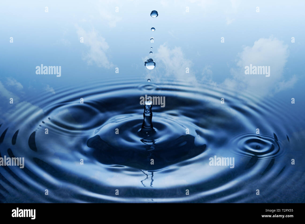 Splash. Round rain drops falling on smooth surface of water. Blue sky and white clouds in background. Stock Photo