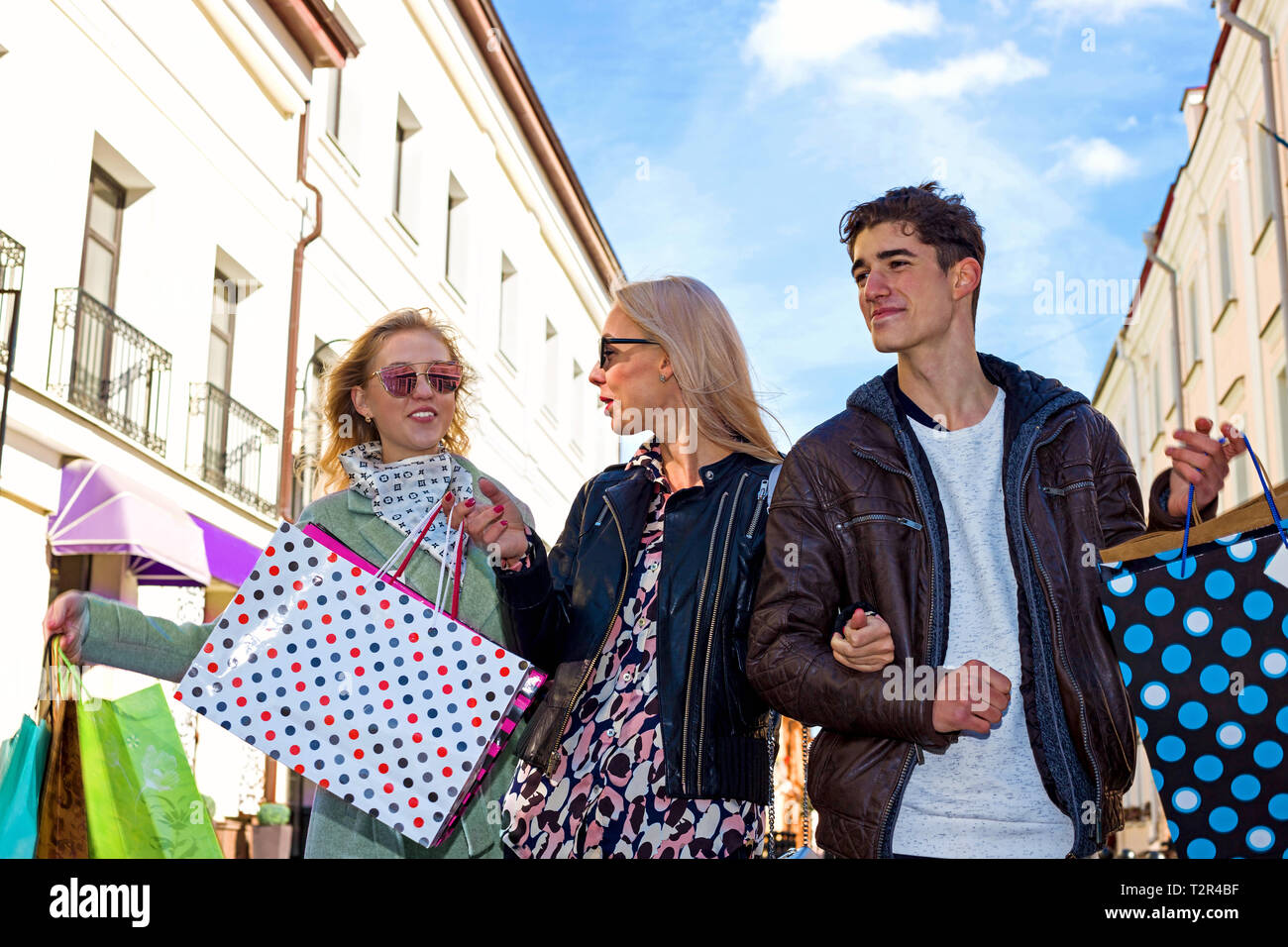 Happy Young two business girls blondes in glasses and a brunet guy walk down the street and enjoy shopping, shopping bags, gifts in their hands. Consu Stock Photo