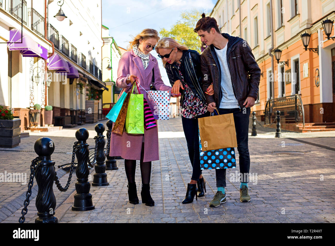 Happy Young two business girls blondes in glasses and a brunet guy walk down the street and enjoy shopping, shopping bags, gifts in their hands. Consu Stock Photo