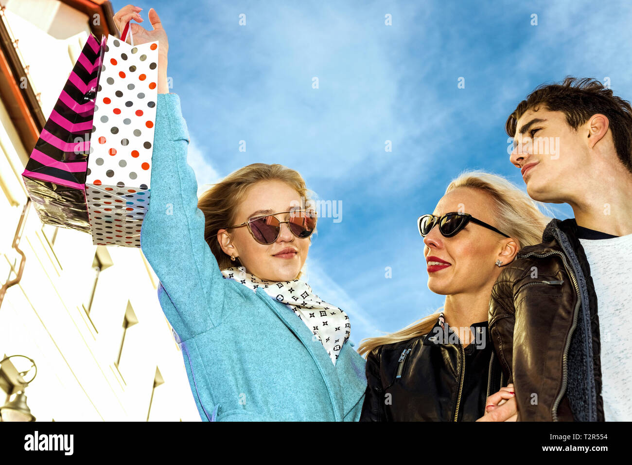 Happy Young two business girls blondes in glasses and a brunet guy walk down the street and enjoy shopping, shopping bags, gifts in their hands. Consu Stock Photo