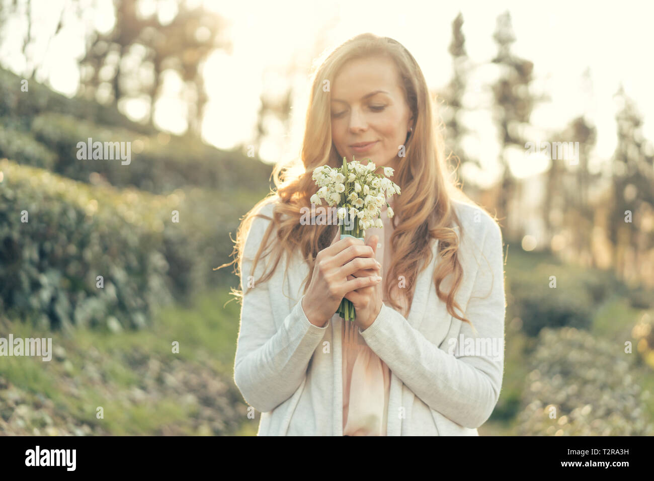 Happy woman with red long hair and a pleasant smile in the rays of the sun holds a bouquet of delicate flowers in front of her and enjoys Stock Photo