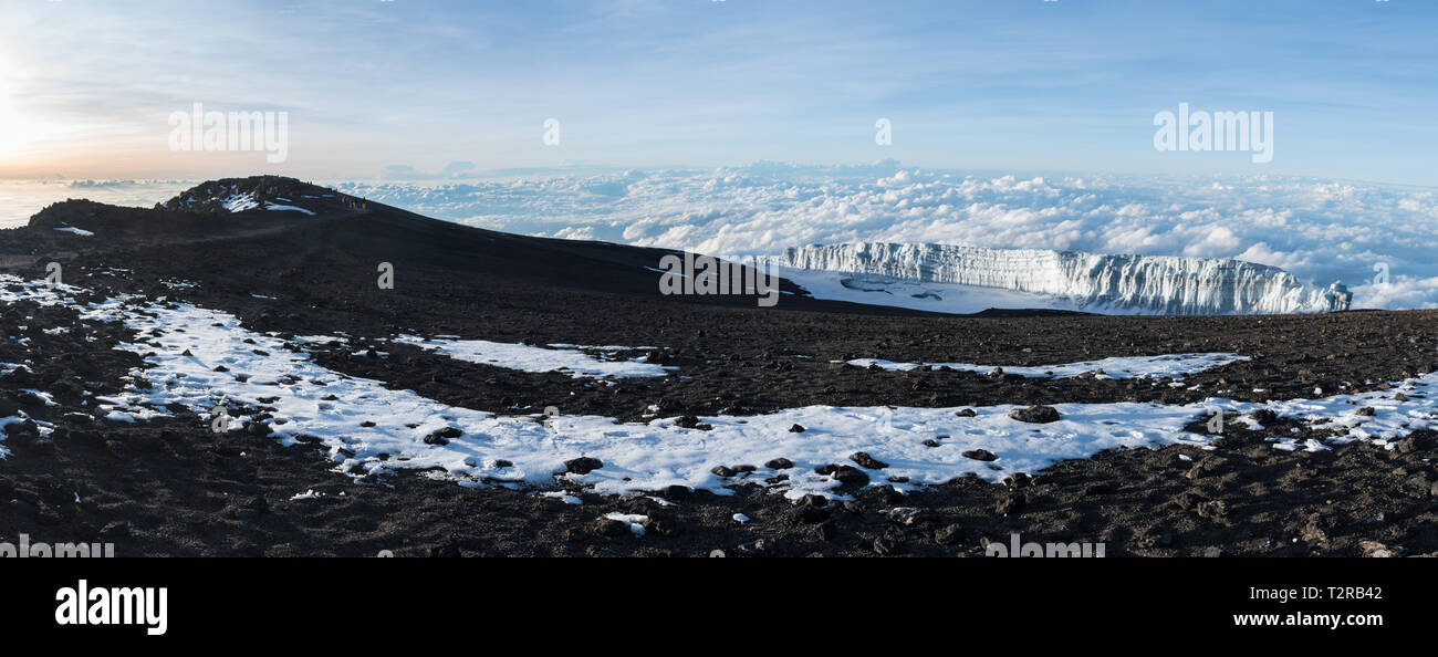 Panoramic view of a glacier sitting above the clouds at the summit of Mount Kilimanjaro taken at sunrise, with unidentifiable people climbing the path Stock Photo