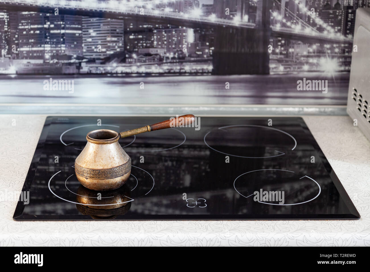 Old Bronze Turkish retro coffee maker kanaka on glass hob and stove with wooden handle, worn on a black background in an apartment in the kitchen. Ret Stock Photo