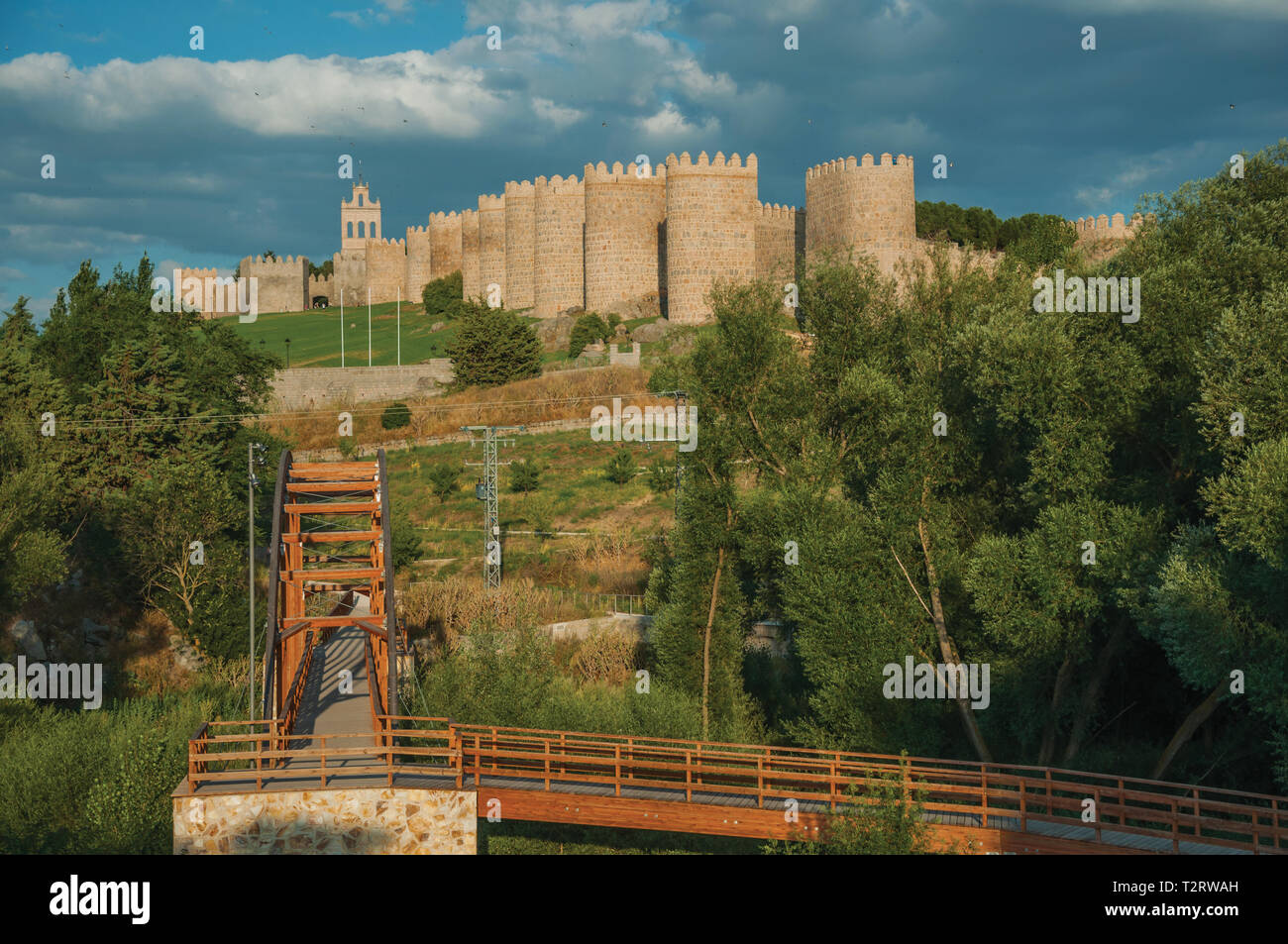 Wooden bridge over the Adaja River with trees and the city of Avila at sunset. With an imposing wall around the gothic city center in Spain. Stock Photo