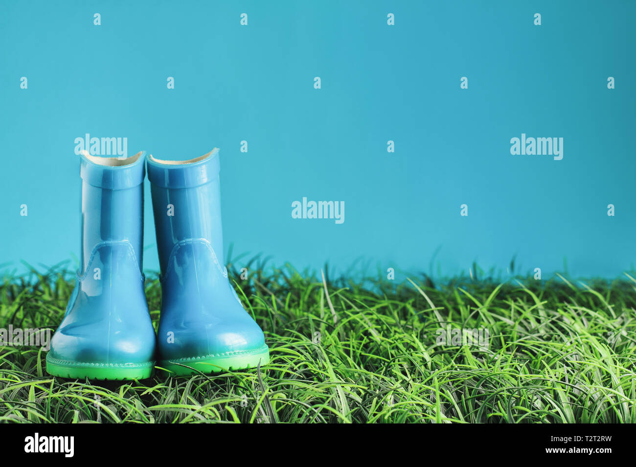 Blue children's rain boots / wellies sitting in the grasss agaisnt a blue background with room for copy space. Stock Photo