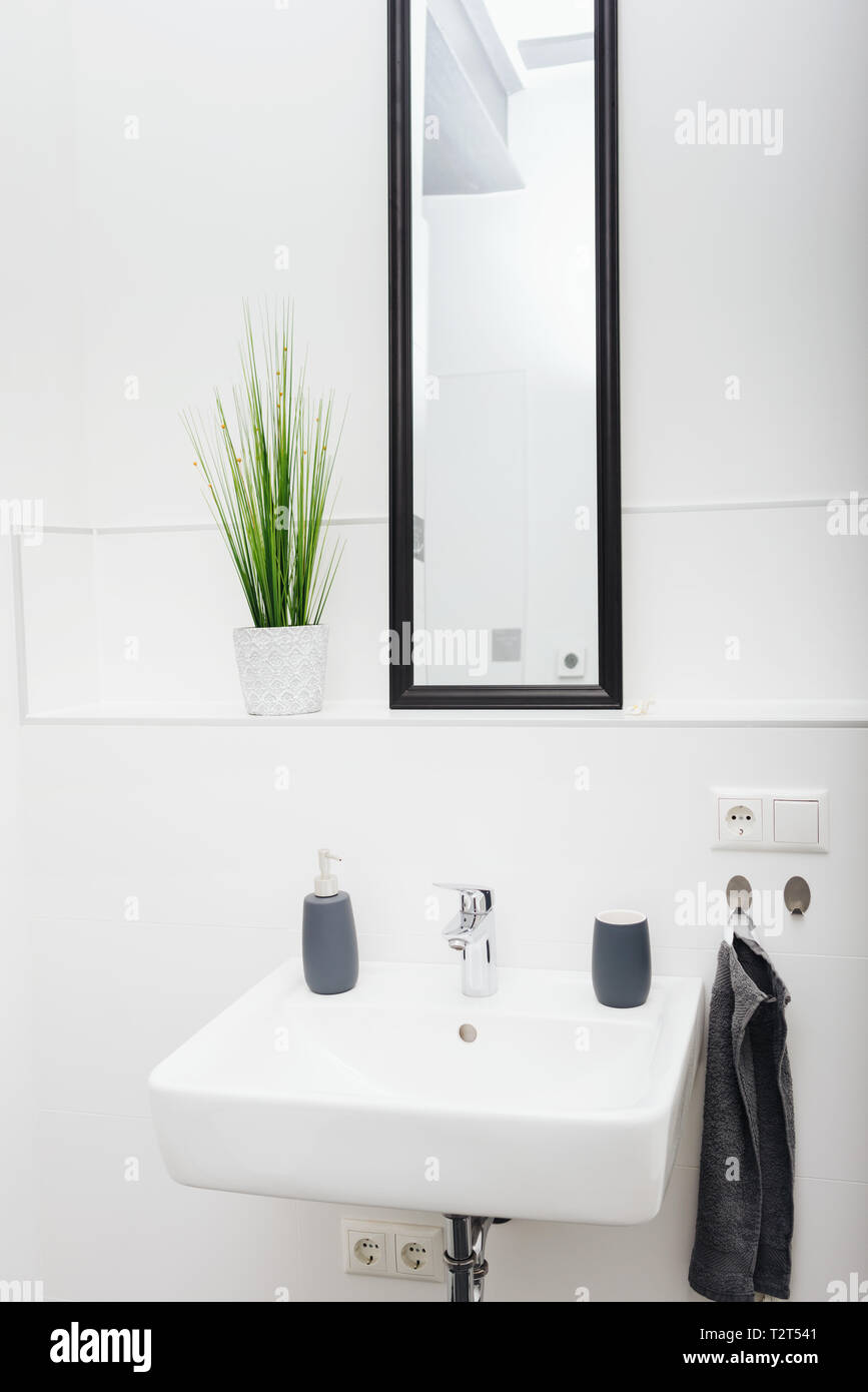 Long tall mirror above a ceramic hand basin in a neat white bathroom with green potted plant and towel on the wall Stock Photo