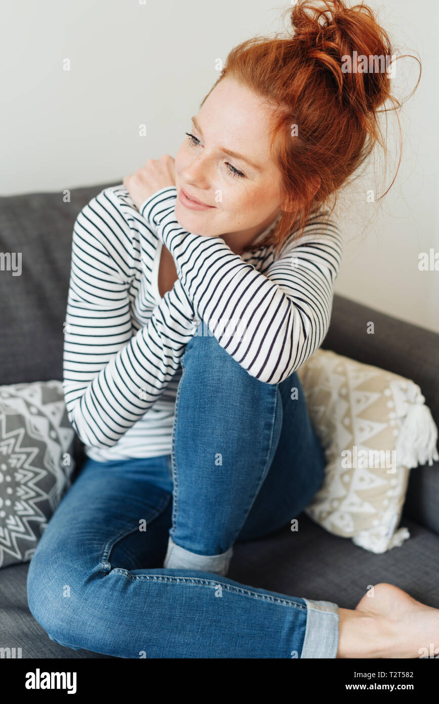 Attractive young redhead woman in denim jeans and striped top sitting  thinking with her legs up on a couch looking aside with a smile Stock Photo  - Alamy