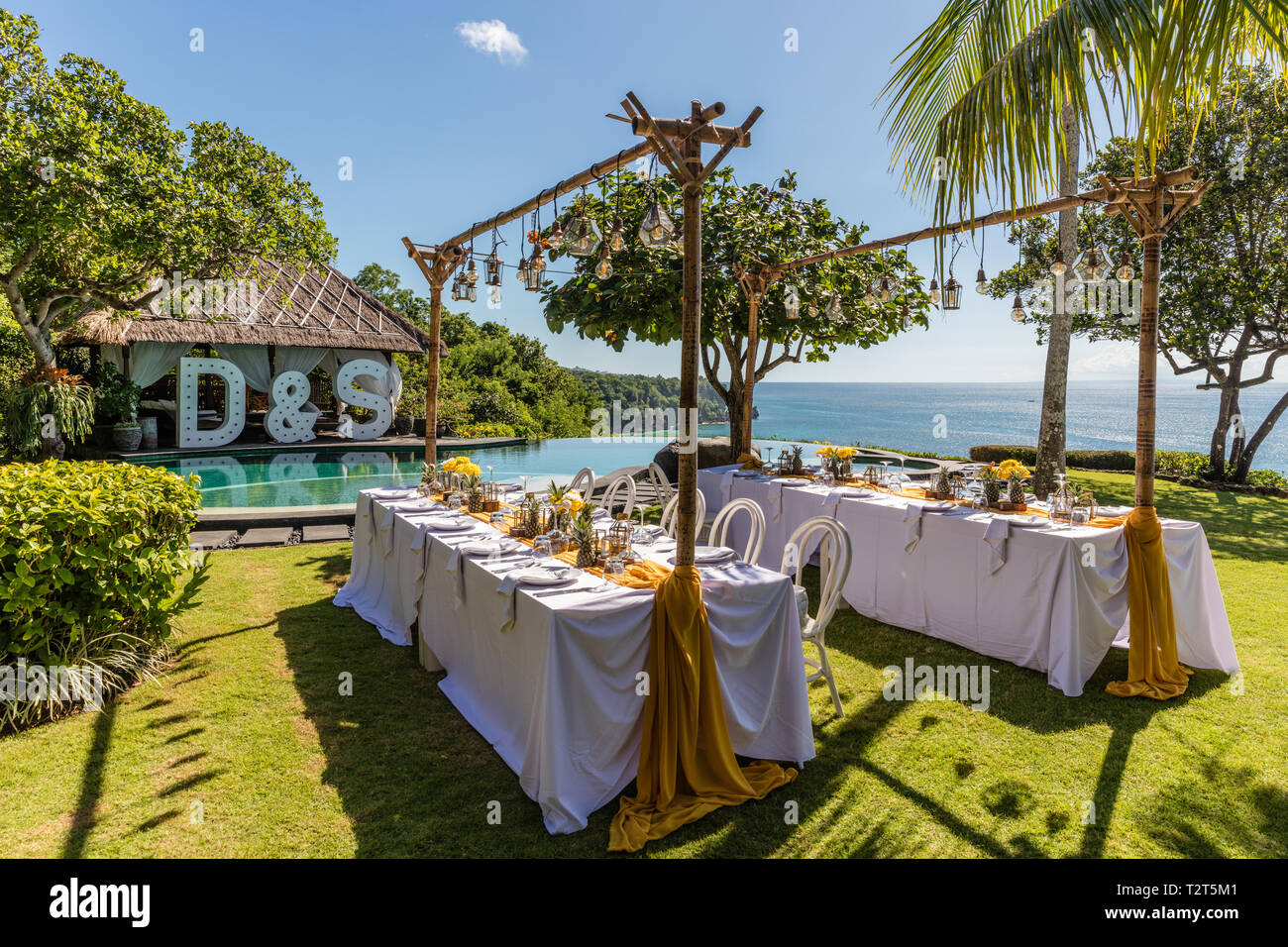 Set white tables for wedding dinner decorated with yellow roses, chrysanthemums, pineapples and glass lamps. View of the ocean. Destination wedding. Stock Photo