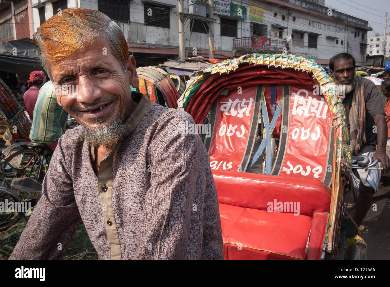 Smiling, happy rickshaw driver with wrinkled face and orange red henna hair on busy Dhaka street, stuck in the traffic jam Stock Photo
