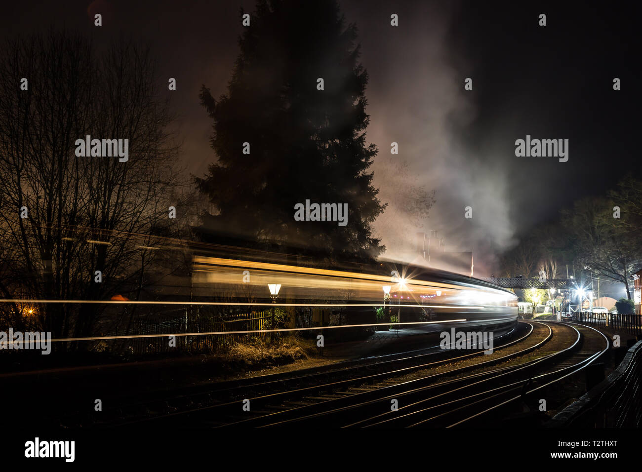 Atmospheric moving steam train & carriages travelling through vintage railway station at night in the dark. Fast movement motion blur. Concept speed. Stock Photo